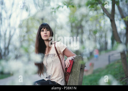 Attraktive junge Frau auf einer Bank im Park sitzen. Kaukasische Mädchen mit langem braunem Haar in einer heiteren darstellen. Stockfoto