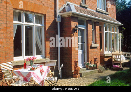 Weiß Klappbare Stühle und Tisch mit rot+weiss Tuch auf Terrasse vor der roten Backstein viktorianischen Landhaus Stockfoto