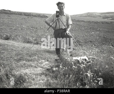 1950er Jahre, begeisterter Amateur Fotograf... ein Mann im t-shirt Ärmel standin auf Gras am Rande eines Landes Feld mit seinem Film Kamera auf einem Stativ, England, UK. Stative werden verwendet, um die Kamera zu stablise. Stockfoto
