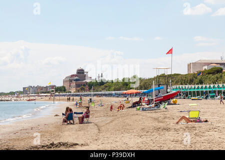 Rettungsschwimmer und Sonnenanbeter am Strand mit Cabanas am Strand oder Hütten, Lido di Venezia, Lido, Venedig, Venetien, Italien. Blick auf Hotel Excelsior Stockfoto