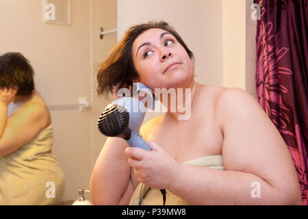 Schöne Frau dreht ihr kurzes Haar, während Sie mit einem Haartrockner, Augen am Morgen ein Lächeln auf den Lippen. Stockfoto