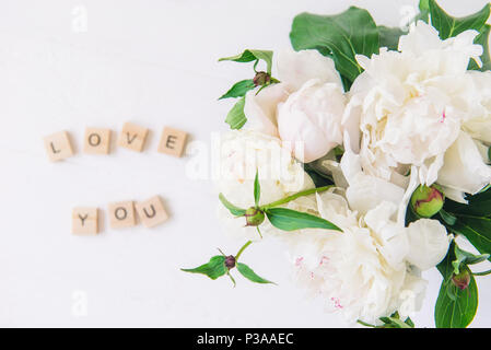 Flach Schwerpunkt auf frischen Blumenstrauß aus den weißen Pfingstrosen und verschwommen Liebe dich Schriftzug in der hölzernen Blöcke geschrieben am hellen Hintergrund. Liebe Worte Konzept. Vale Stockfoto