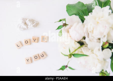 Flach Liebe dich Schriftzug in Holzblöcke mit statuette von zwei antike wunderschöne Engel mit unscharfen weiße Pfingstrose auf hellen Hintergrund geschrieben. Liebe Stockfoto