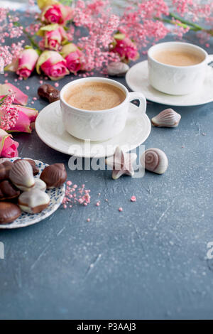 Eine Tasse duftenden Kaffee am Morgen und ein Blumenstrauß rosa Blüten. Süßigkeiten Schokolade in Form von cockleshells. Muscheln an den Cups. Romantik und hellen Farben. Guten Morgen. Postkarte. Für tex Platz Stockfoto