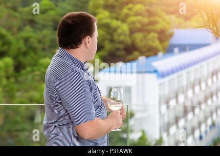 Junge Unternehmer in Freizeitkleidung auf transparentem Glas Balkon und Blick auf die City Resort. Mann hält ein Glas Weißwein und Entspannen genießen Stockfoto