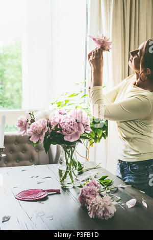 Hübsche Frau mit rosa Blumen Pfingstrosen am Tisch im Wohnzimmer, und Strauß in einem Glas Vase am Fenster. Sommer noch leben. Gemütlich zu Hause Szene Stockfoto