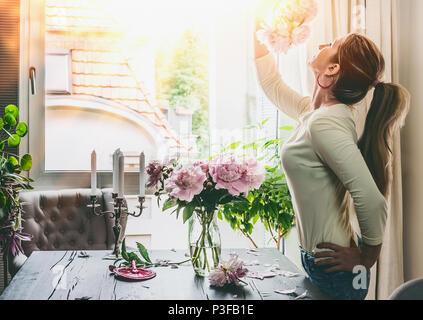 Schöne Frau mit rosa Blumen Pfingstrosen auf dem Tisch im Wohnzimmer komponiert ein Blumenstrauß in einem Glas Vase am Fenster. Sommer noch leben. Gemütlich zu Hause s Stockfoto