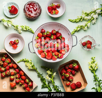 Frische Erdbeeren in Sieb, Marmelade im Glas Glas mit Sommer Blumen und frischen Beeren in Box, Schüsseln und Löffel auf dem Tisch Hintergrund, Ansicht von oben. Beeren Stockfoto