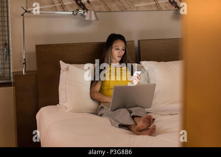 Geschäftsfrau, sitzen auf dem Bett können Sie über Ihr Telefon während der Arbeit am Laptop Stockfoto