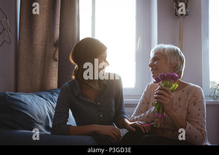 Tochter geben ältere Frau Blumen beim Sitzen auf einem Sofa im Wohnzimmer. Stockfoto