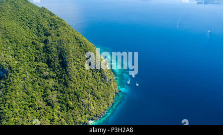 Antenne drone Blick auf die Boote im klaren Meer, über ein tropisches Korallenriff, umgeben von aufragenden Dschungel bedeckten Berge Stockfoto