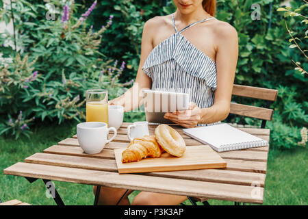 Junge und attraktive Frau mit Frühstück im grünen Garten mit französischen Croissants, Donuts, Kaffee, Orangensaft, Tablet und Hinweise Buch über Stockfoto