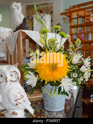 Ein helles Orange Blume steht im Mittelpunkt einer Blume Anordnung in einem verzinkten Zinn Vase. Dekorative Eule Statuen und andere Objekte werden auch dargestellt. Stockfoto