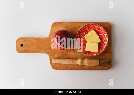 Ein roter Apfel, eine kleine Platte mit Butter und ein backblech Pinsel auf einer hölzernen Schneidebrett mit einem weißen Tag. Zutaten für einen Apfelkuchen. Essen Konzept. Stockfoto