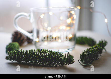 Nahaufnahme einer unscharfen Glas Becher mit warmen Weihnachtsbeleuchtung innen durch Tannenzapfen und Zweige auf einen weißen Tisch umgeben. Weihnachten Konzept. Stockfoto