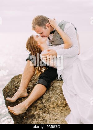 Die outdoor Portrait des Küssens Jungvermählten. Der Bräutigam sitzt auf der Klippe während die Braut auf seine Runden auf dem Hintergrund des Meeres liegt. Stockfoto