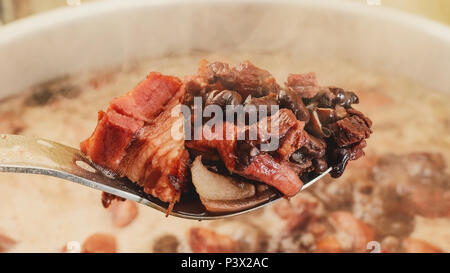 Löffel aus der Pfanne Zutaten der Feijoada, brasilianischer Küche. Löffel mit schwarzen Bohnen, Schweinefleisch und Speck. Feijoada Kochen in den Topf auf. Stockfoto