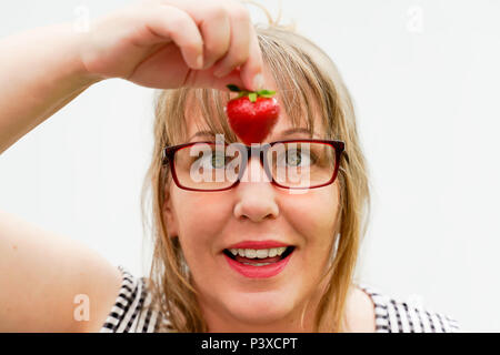 Eine Frau mittleren Alters hält eine Erdbeere. Sie hat blonde Haare und trägt eine Brille. Ihre Augen blick in die Erdbeere, so Schiele. Stockfoto