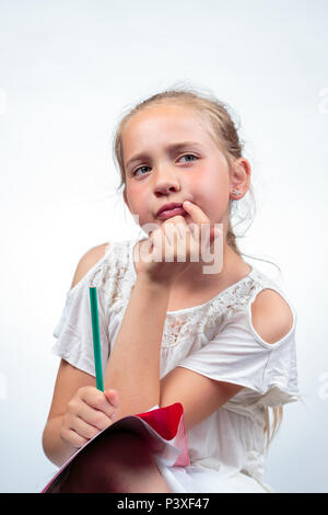 Eine hübsche 10-jährige kaukasischen Schulmädchen denken ruht Ihr Gesicht in ihre Hand, während ein Bleistift und Ihre Notizen auf ihrem Knie gegen ein Licht zurück Stockfoto