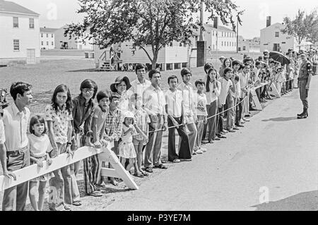 FORT SMITH, AR, USA - 10. AUGUST 1975 - eine militärische Polizist beobachtet, wie eine Linie der neu angekommenen Vietnamesische Flüchtlinge stehen und für die Weitergabe von Präsident Gerald Ford warten. Der Präsident ist zu touring das Fort Chaffee vietnamesischer Flüchtling Zentrum sein und einladend Die evakuierten Südvietnamesischen in die Vereinigten Staaten. Hinter den Menschen steht die Zeilen der Kasernengebäude, dass die Flüchtlinge untergebracht. Stockfoto