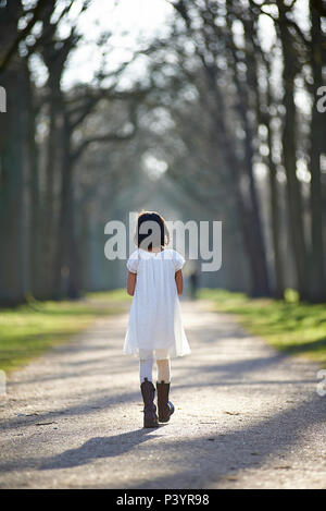 Süße asiatische Mädchen in einem weißen Kleid zu Fuß auf einen Weg in die Perspektive in einem Park in der schönen Frühlingssonne Stockfoto
