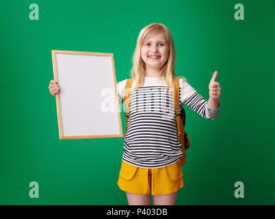 Lächelnd Schüler mit Rucksack mit Daumen nach oben und und Blank Board gegen grüner Hintergrund Stockfoto