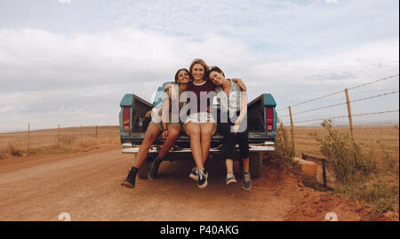 Portrait von drei jungen weiblichen Freunde an der Rückseite der Pickup Truck auf einer Landstraße geparkt. Gruppe von Frauen auf dem Land road trip. Stockfoto