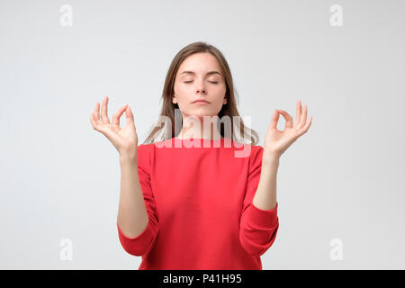 Indoor Portrait von ruhigen schönen kaukasischen Frau Meditation in rotes Hemd, lächelnd, während die Hände mit Zen Gesten, stehend gegen Grau backgr Stockfoto