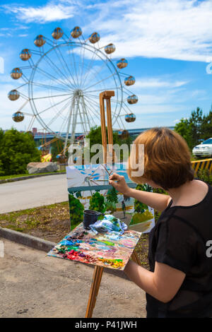 Eine blonde junge Künstlerin in einem schwarzen T-Shirt und Jeans Farben auf Leinwand und einer Staffelei mit Ölfarben in einem Stadtpark an einem sonnigen Tag. Zeichnung auf einer o Stockfoto