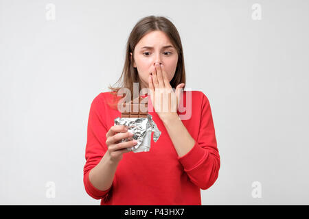 Portrait traurige junge Frau in Rot Pullover müde von Ernährung. Sie möchten Schokolade auf grauen Hintergrund isoliert zu essen. Menschliches Gesicht Ausdruck Stockfoto