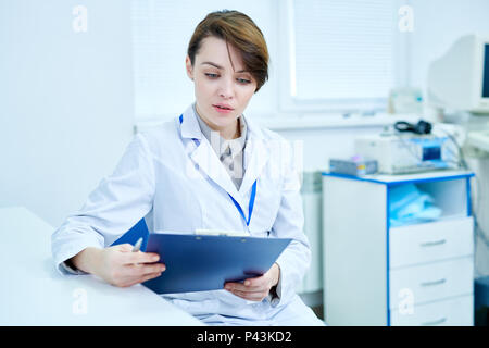 Arzt sitzen am Schreibtisch holding Zwischenablage und Lesen Stockfoto