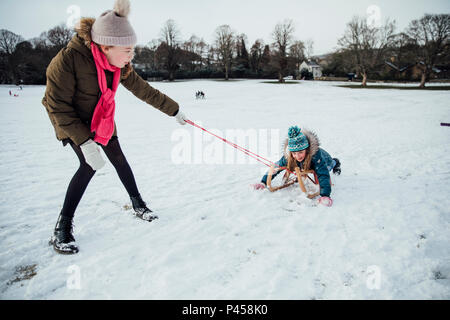 Zwei kleine Mädchen haben Spaß im Schnee. Eines der Mädchen Zieht die andere auf einem Schlitten. Stockfoto
