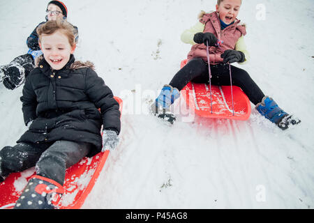 Kinder aus der Gemeinschaft sind auf einem Hügel in einem öffentlichen Park auf Schlitten in den Schnee. Stockfoto