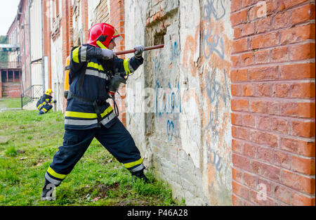 Ansicht von hinten auf Feuerwehrmann in Uniform mit voller Schutzausrüstung, mit Brecheisen die Mauer durchbrechen. Stockfoto