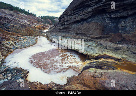 Strom von Acid orange Wasser mit einem niedrigen PH-Wert zwischen den Felsen mit schwarzen und roten Felsen, mit Pinienwald im Hintergrund in der alten Zaranda Mine, Sp Stockfoto