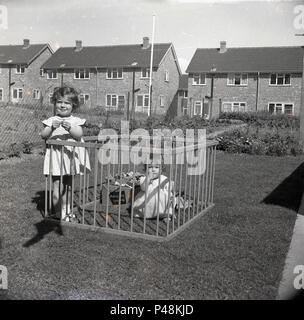 1950, historische, Baby Mädchen sitzen in ihrem Holz Laufgitter draußen auf dem Rasen im Garten, mit ihrer älteren Schwester auch stehend in es, England, UK. Stockfoto