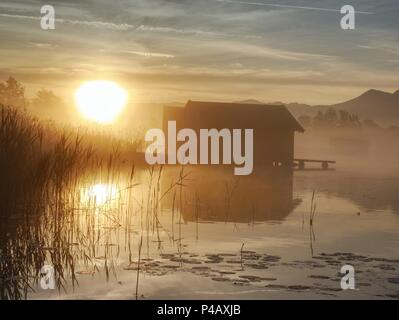 Romantischen sonnenaufgang Reflexion auf dem Kochelsee gegen Bootshäusern. Anfang Sommer morgen ansehen, Bayern, Deutschland Stockfoto