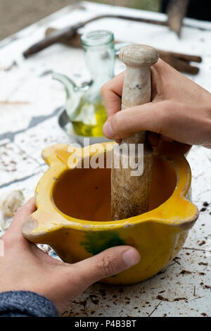 Nahaufnahme von einem kaukasischen Mann Vorbereitung ein hausgemachter Sauce oder Schleifen einige Zutaten in einem alten Mörser aus Keramik, mit einem hölzernen Stampfe, auf einem rustikalen Tisch o Stockfoto