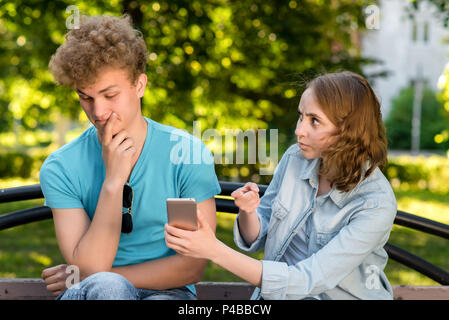 Ein junges Mädchen zeigt mit dem Finger auf Smartphone Kerl. Das Problem in der Beziehung. Gefühle des Misstrauens. Konzepte von Verrat und Verrat. Junges Paar im Sommer in einem Park im Freien. Stockfoto