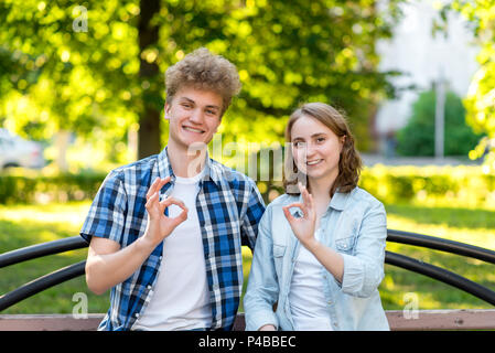 Ein Kerl mit einem Mädchen im Sommer in einem Park in der Natur. Lächeln emotional zufrieden. Geste zeigt auf OK. Das Konzept der Erfolg und viel Glück. Glücklich die Schüler nach der Schule. Stockfoto