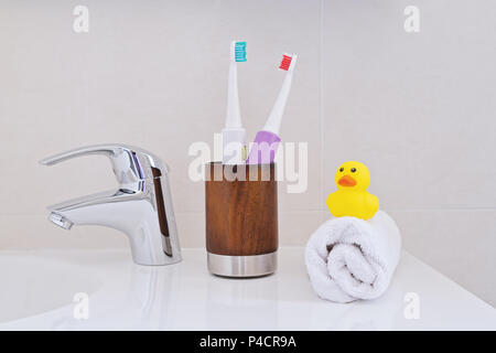 Zwei elektrische Zahnbürsten in Holz Glas am Waschbecken im Badezimmer mit weißen Handtuch eingewickelt und gelb Ducky Duck. Persönliche Hygiene Konzept. Stockfoto