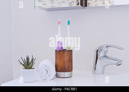 Zwei elektrische Zahnbürsten in Holz Glas am Waschbecken im Badezimmer mit weißen Handtuch gewickelt. Chrom Wasserhahn und Waschbecken. Bad Interieur und Zubehör Stockfoto