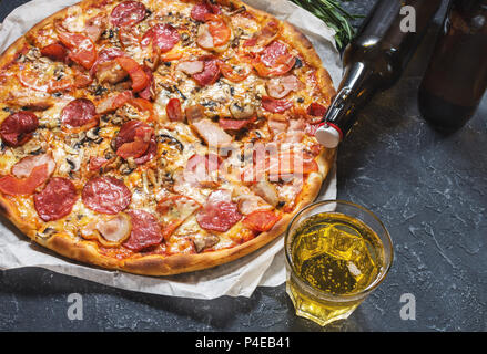 Frische, hausgemachte Pizza mit Salami, Schinken, Käse und ein Glas Bier auf rustikalem Stein Hintergrund. Stockfoto