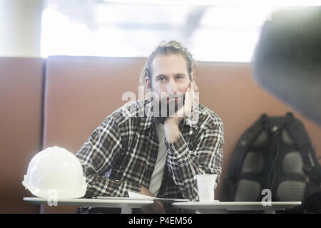 Mann mit seinen Kopf in seinen Händen an einem Tisch sitzen mit einem Schutzhelm neben ihm Stockfoto