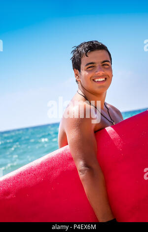 Closep Portrait Of Happy fröhlich surfboarder mit roten Surfbrett stehend über blauer Himmel, seinem Hobby, aktiven Lebensstil der Jugend Stockfoto