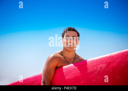 Portrait Of Happy fröhlich surfboarder mit roten Surfbrett stehend über blauer Himmel, seinem Hobby, aktiven Lebensstil der Jugend Stockfoto