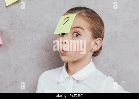 Portrait von kleinen Mädchen mit Haftnotiz mit Fragezeichen auf dem Gesicht isoliert auf Grau Stockfoto