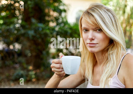 Fröhliche junge Frau mit langen blonden Haare im Licht Sommer top Holding in der Hand kleine Tasse mit Tee oder Kaffee und mit Blick auf die Kamera lächelnd auf unscharfen Ba Stockfoto