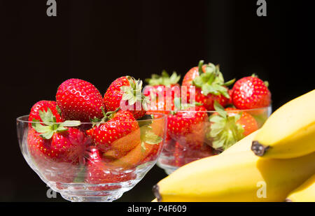 Nahaufnahme von zwei Kristallglasgerichten voller frischer, saftiger Erdbeeren mit Blättern. Ein paar Bananen in Schuss. Schwarzer Hintergrund. Stockfoto