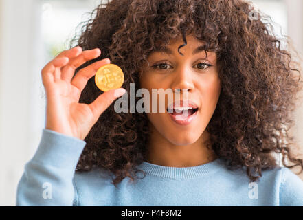 African American woman holding golden Bitcoin cryptocurrency zu Hause Angst im Schlag mit einer Überraschung konfrontiert, ängstlich und aufgeregt mit Angst Ausdruck Stockfoto